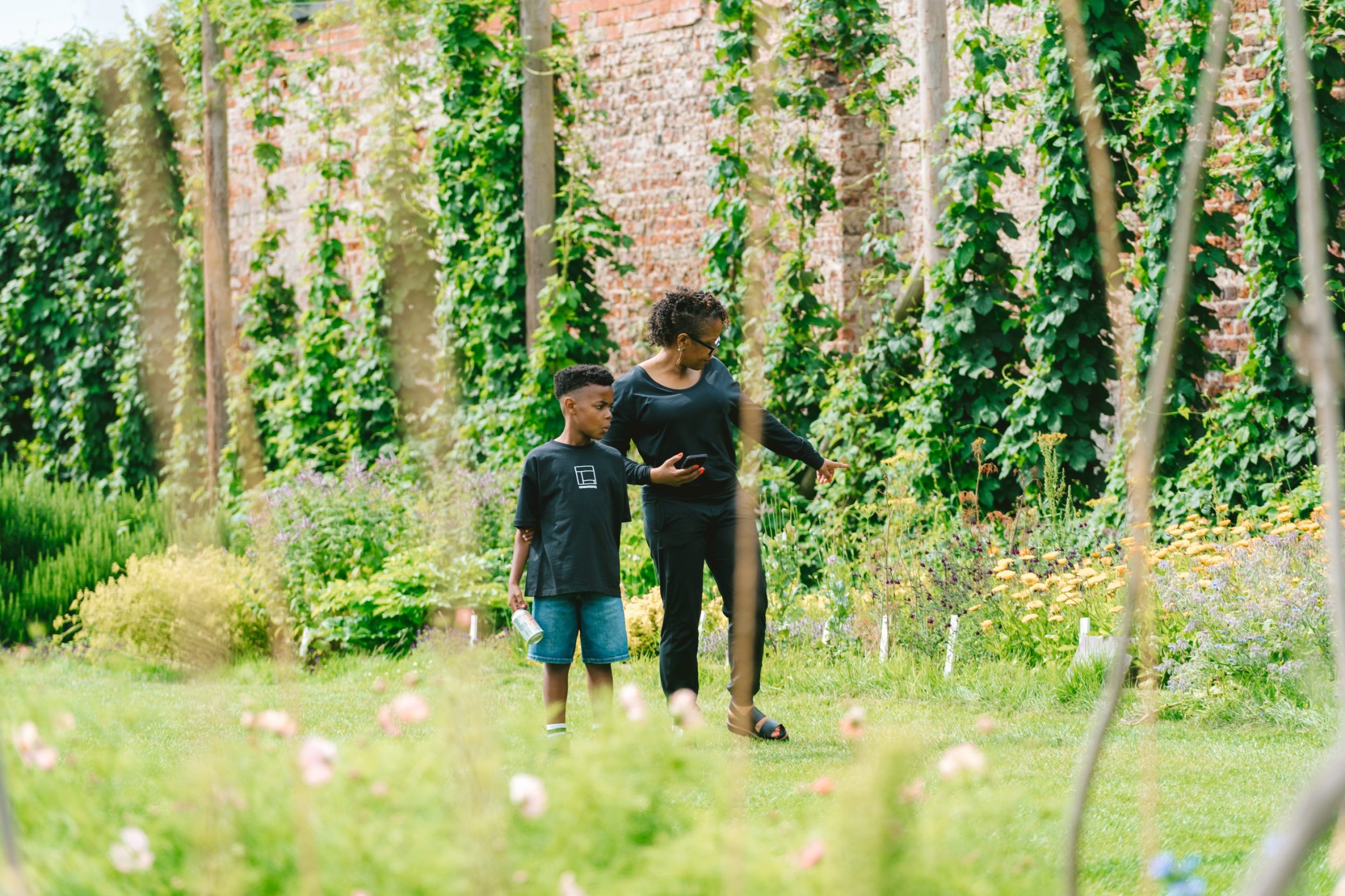 A young boy walks with his mother in a pretty garden, with hops growing up the red brick wall behind them