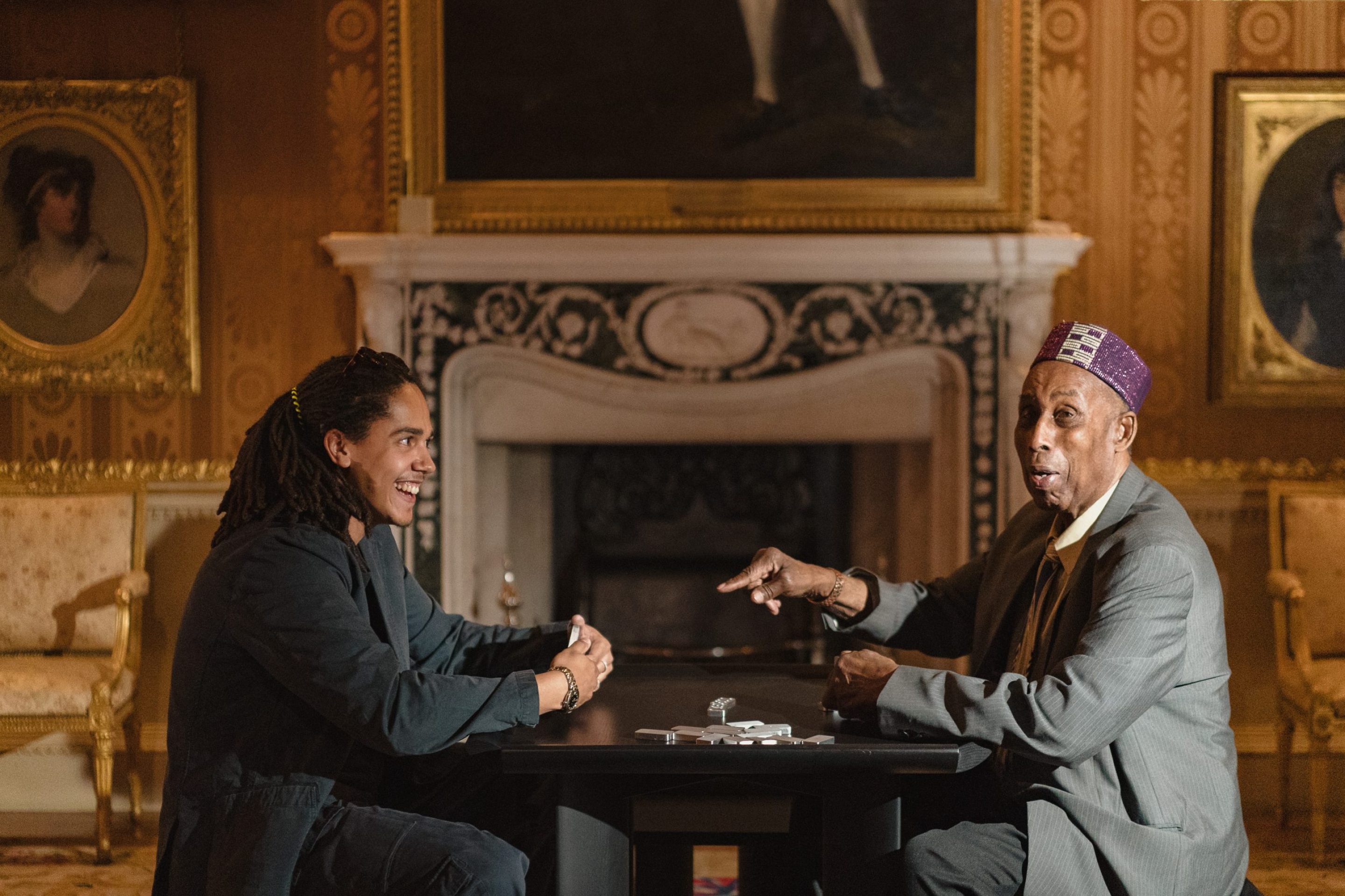 A young Black man with dreadlocks smiles at an older Black man wearing a traditional cap, as they play dominoes in a grand room