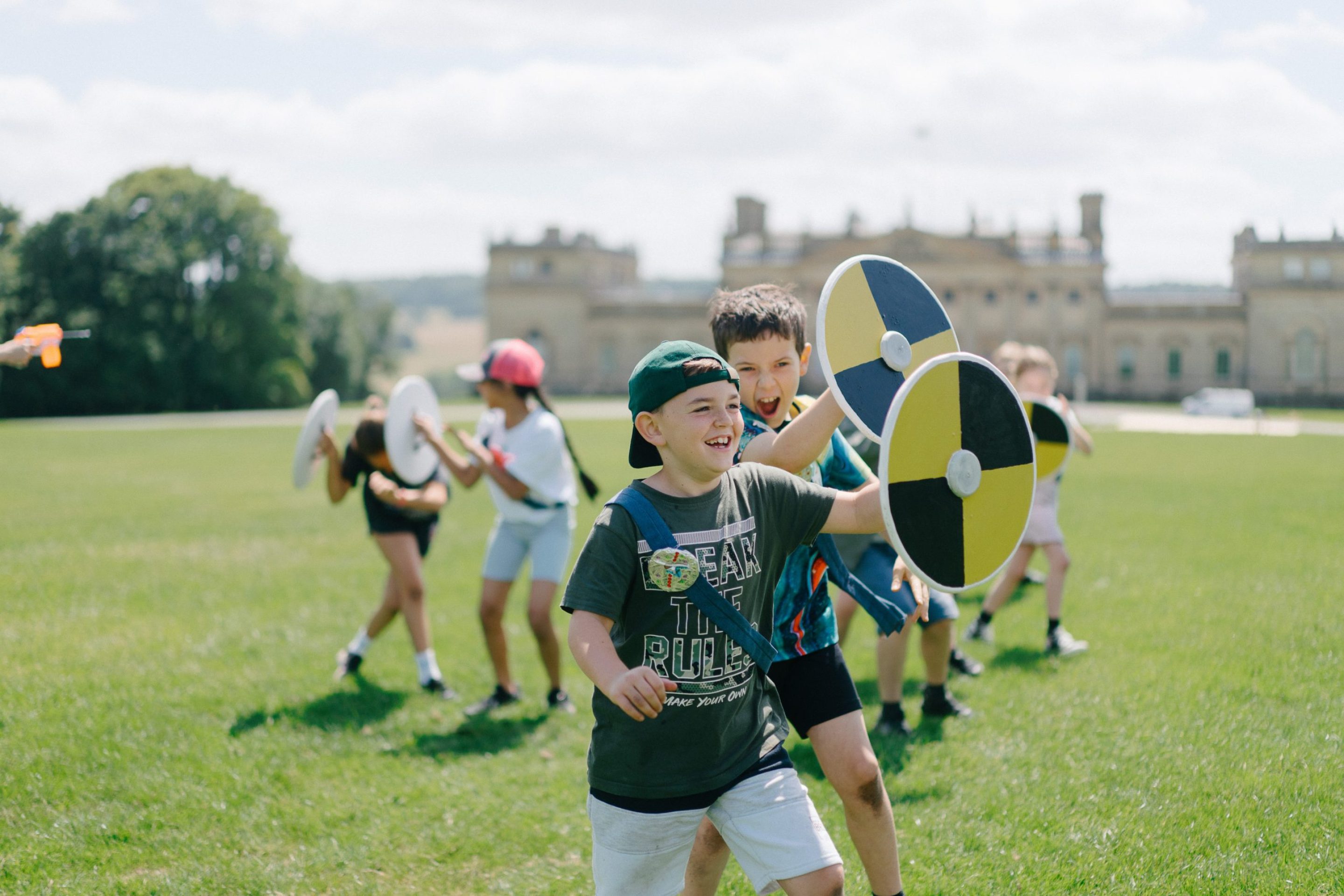 A group of school children playfully wield foam shields on the front lawn of Harewood House. The scene is lively and joyful as they reenact a battle from the Saxon era.