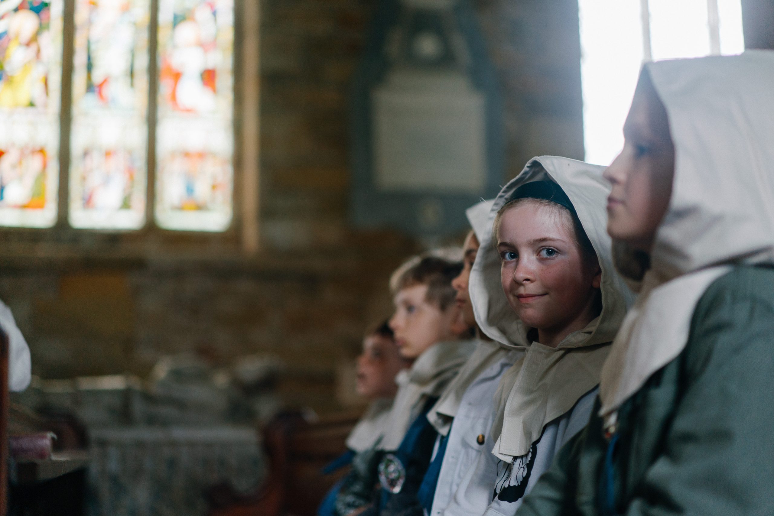 Uploaded image preview A group of children is sitting quietly in a dimly lit church, wearing hooded cloaks. One child, glancing sideways, has a soft smile. Stained glass windows in the background.