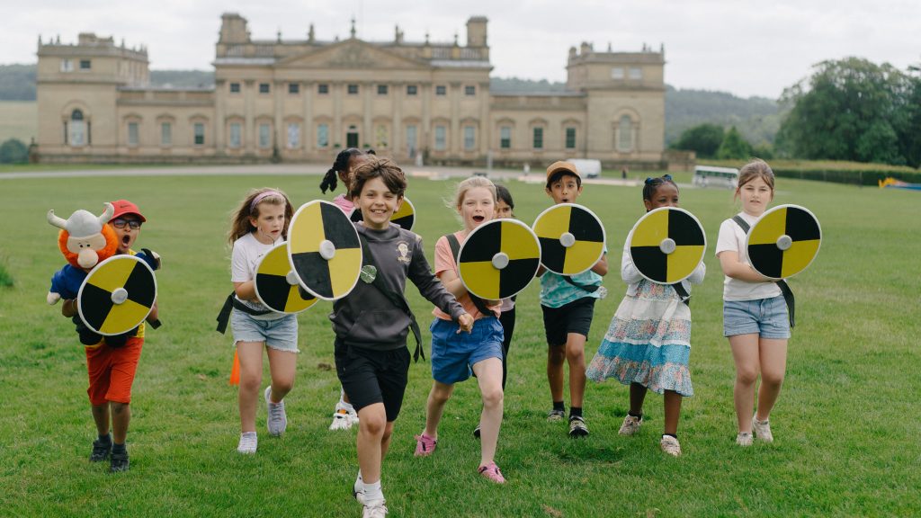 A group of school children playfully wield foam shields on the front lawn of Harewood House. The scene is lively and joyful as they reenact a battle from the Saxon era.