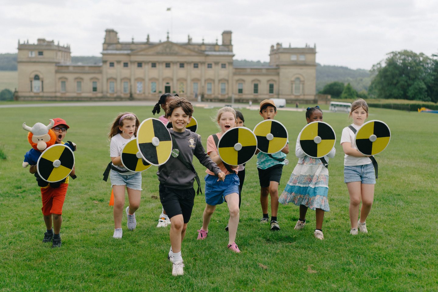 A group of school children playfully wield foam shields on the front lawn of Harewood House. The scene is lively and joyful as they reenact a battle from the Saxon era.