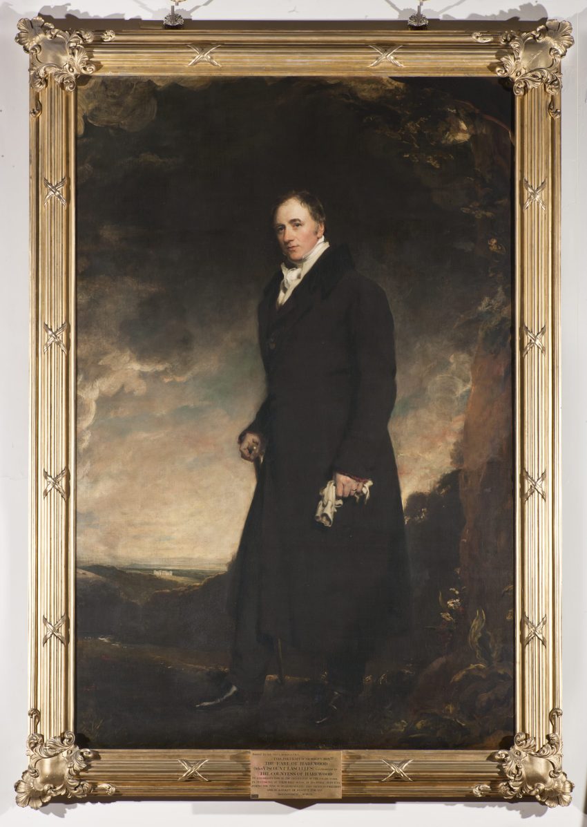 Painted portrait of Henry Lascelles, a middle-aged white man with dark hair, dressed in a long black coat with a wide collar. He is wearing a white shirt with a white neckerchief poking out above his coat, he has a smirk-like expression on his face. He stands turning to the right in front of an expansive landscape. The portrait is housed in a gilt frame.