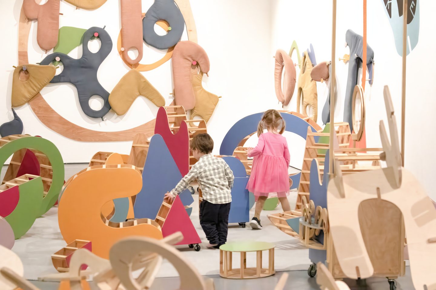 Two children, a boy in a plaid shirt and a girl in a pink dress, explore a colourful, abstract play area with geometric shapes made of soft materials and wood on the walls and ground.