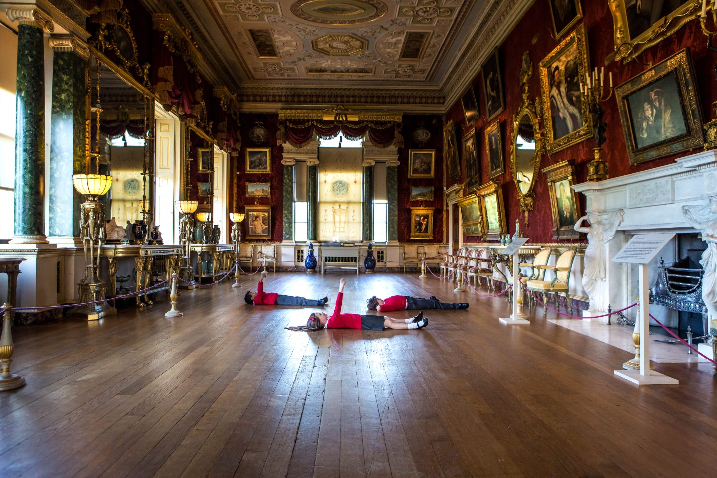 Three young pupils in school uniform lay down in the middle of the long Gallery looking up at the intricate designs of the ceilings. Sunlight beams through the windows, illuminating the golden frames of the many paintings that hang on the opulent red walls.