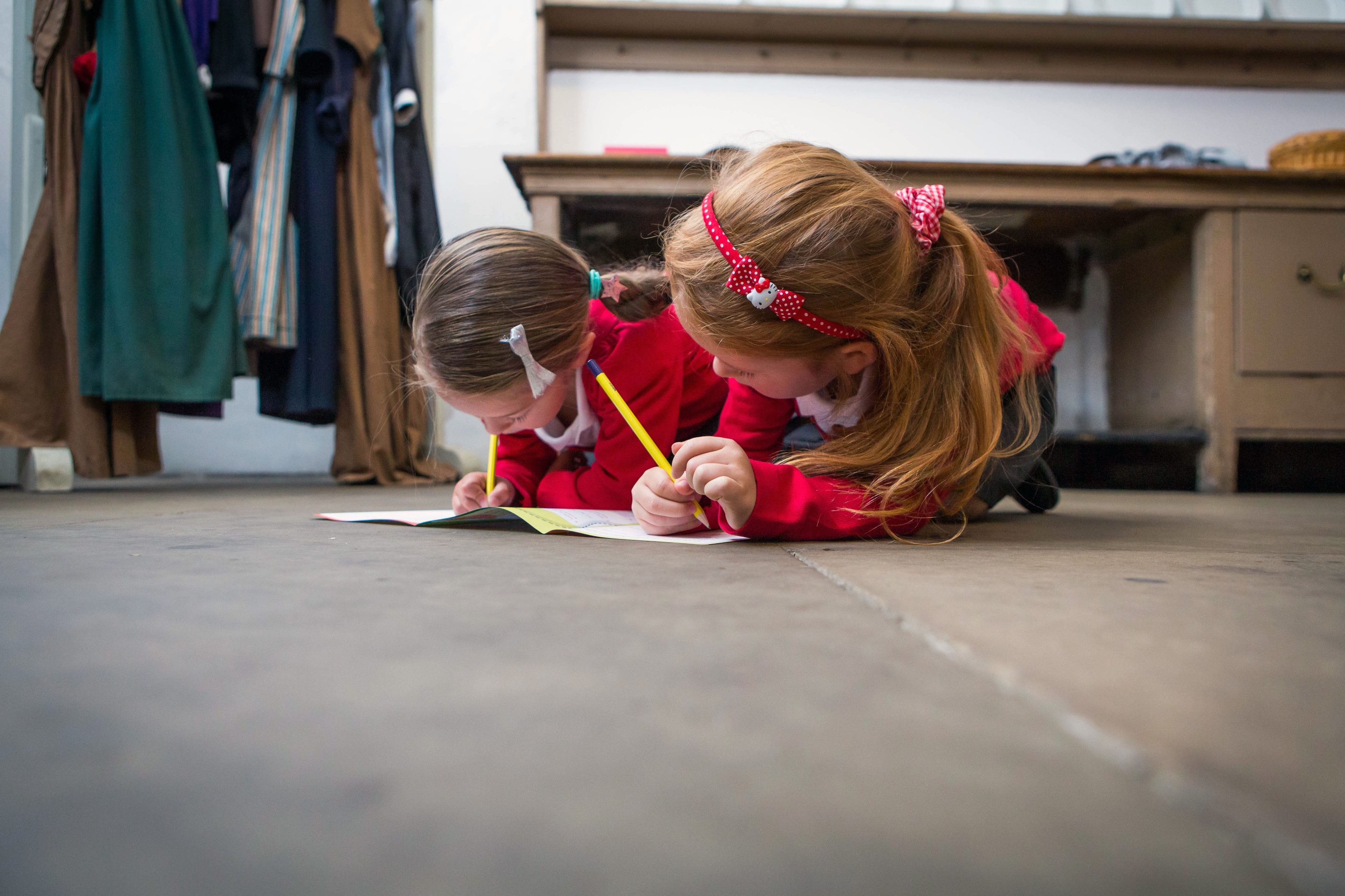 Two young pupils, in red school uniform, kneel on the stone floor to fill out a work book together.