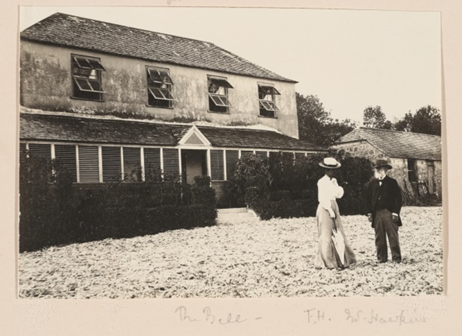 A black and white photo of a man and a woman stood in the foreground in Edwardian-style dress. The woman wears a long skirt, a white puff sleeve shirt, a large hat and holds an umbrella. The man wears a dark tailored jacket and trousers, a wide brimmed hat and has a white beard. There is a large building in the background, with four open windows on the top floor and a front porch below with an opening for the front door.