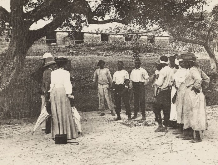 A black and white photograph from 1906 showing a group of Black people in front of a low building, in Barbados