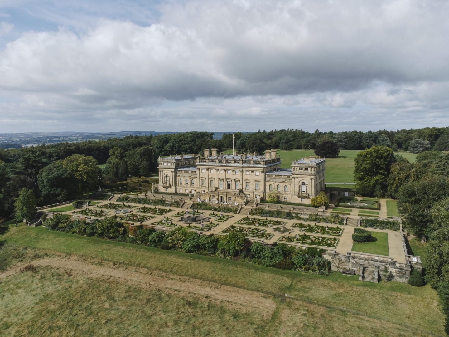 Harewood House, a very large and grand building, is seen from the air, surrounded by manicured gardens with woodlands in the distance