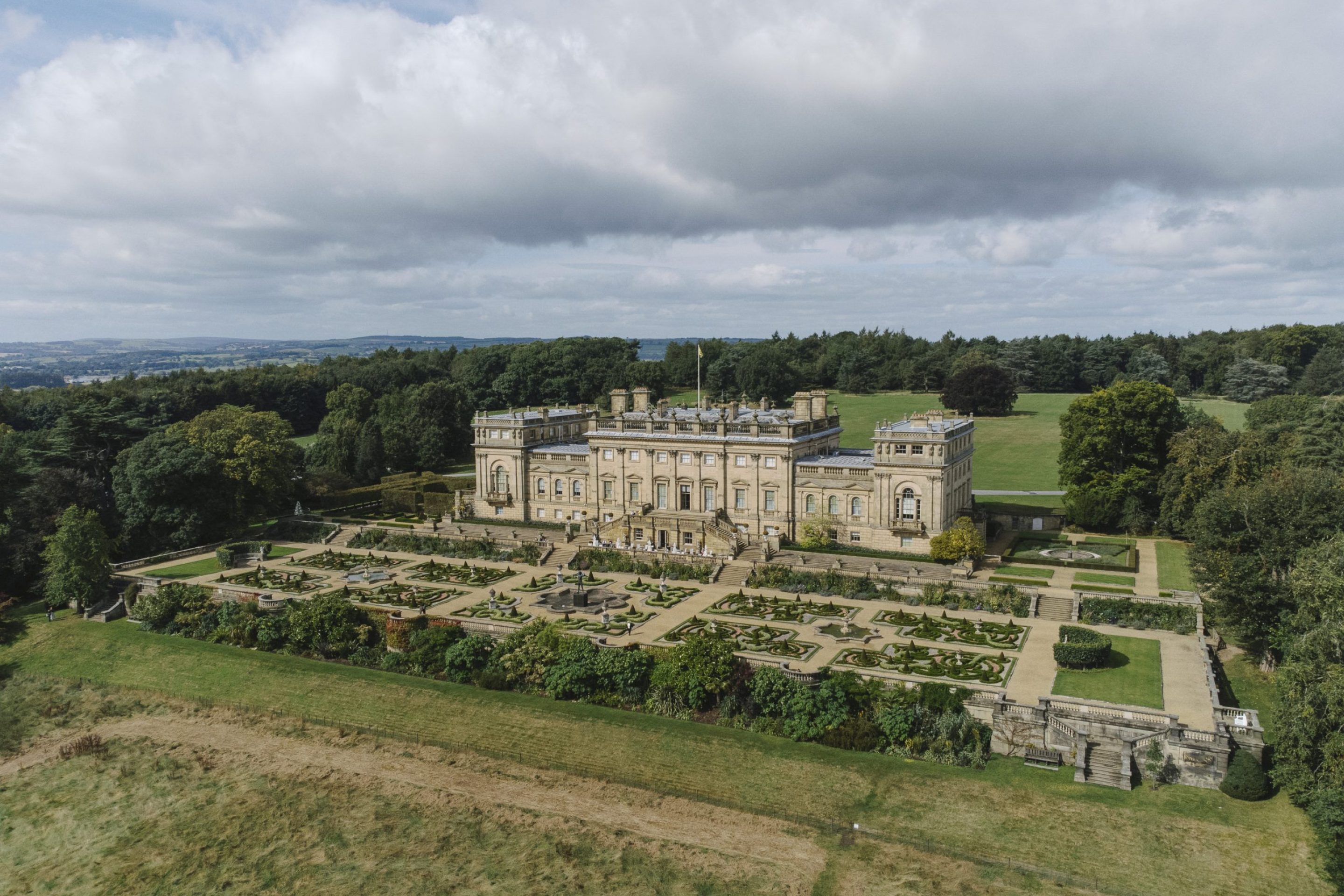 Harewood House, a very large and grand building, is seen from the air, surrounded by manicured gardens with woodlands in the distance