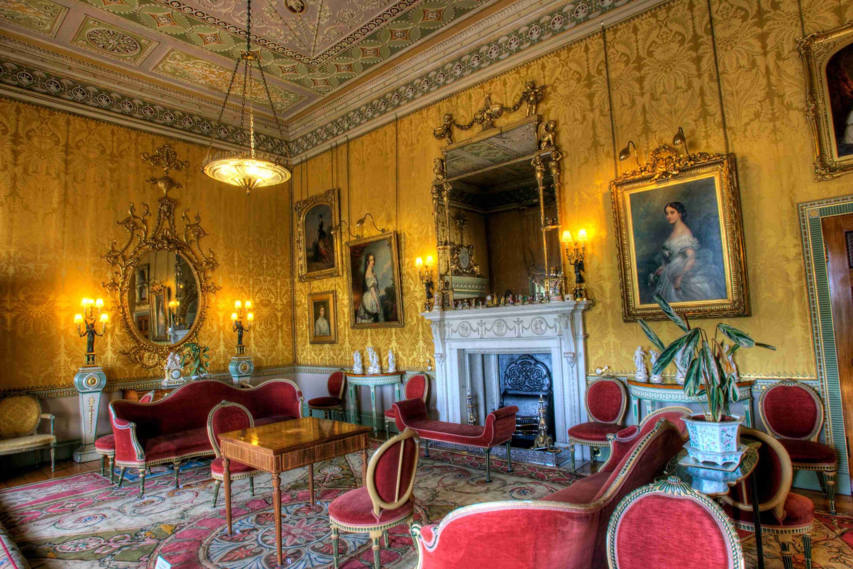 A grand 18th century drawing room with rich Yellow damask walls, decorated with large gilded mirrors and portraits of women. The room is filled with red velvet seat furniture and a large white marble mantlepiece.