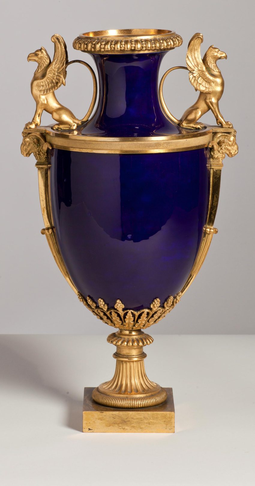 An urn-shaped dark-blue vase encased by gilded metal mounts. Either side of the neck are two small gilded metal gryphons positioned to make handles.